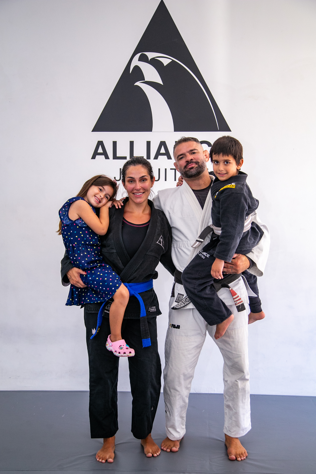 Família de alunos Alliance Viamão em kimono, com crianças, em frente ao logo da academia
