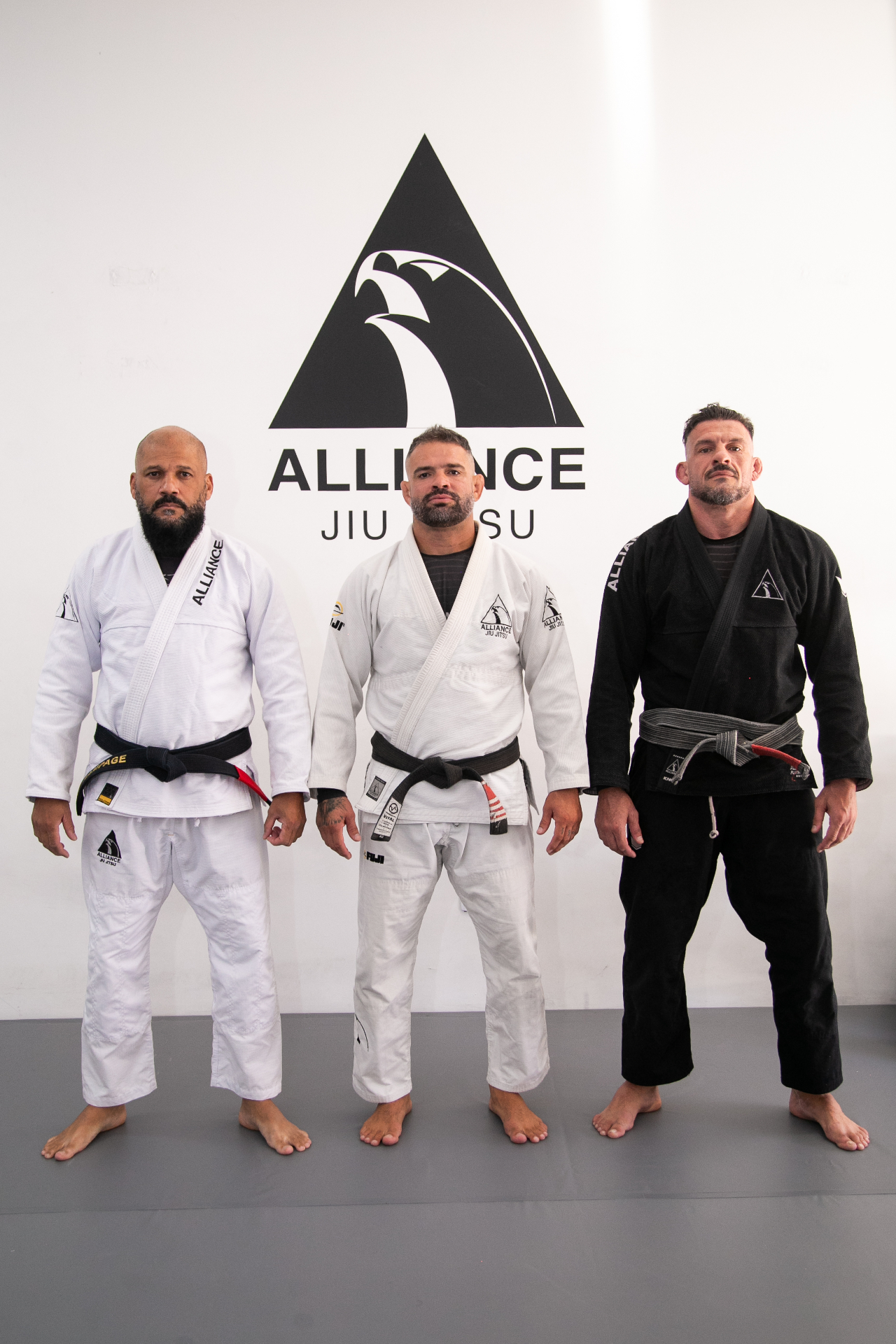 Três professores faixa-preta da Alliance Viamão em pé, em frente ao logo da academia